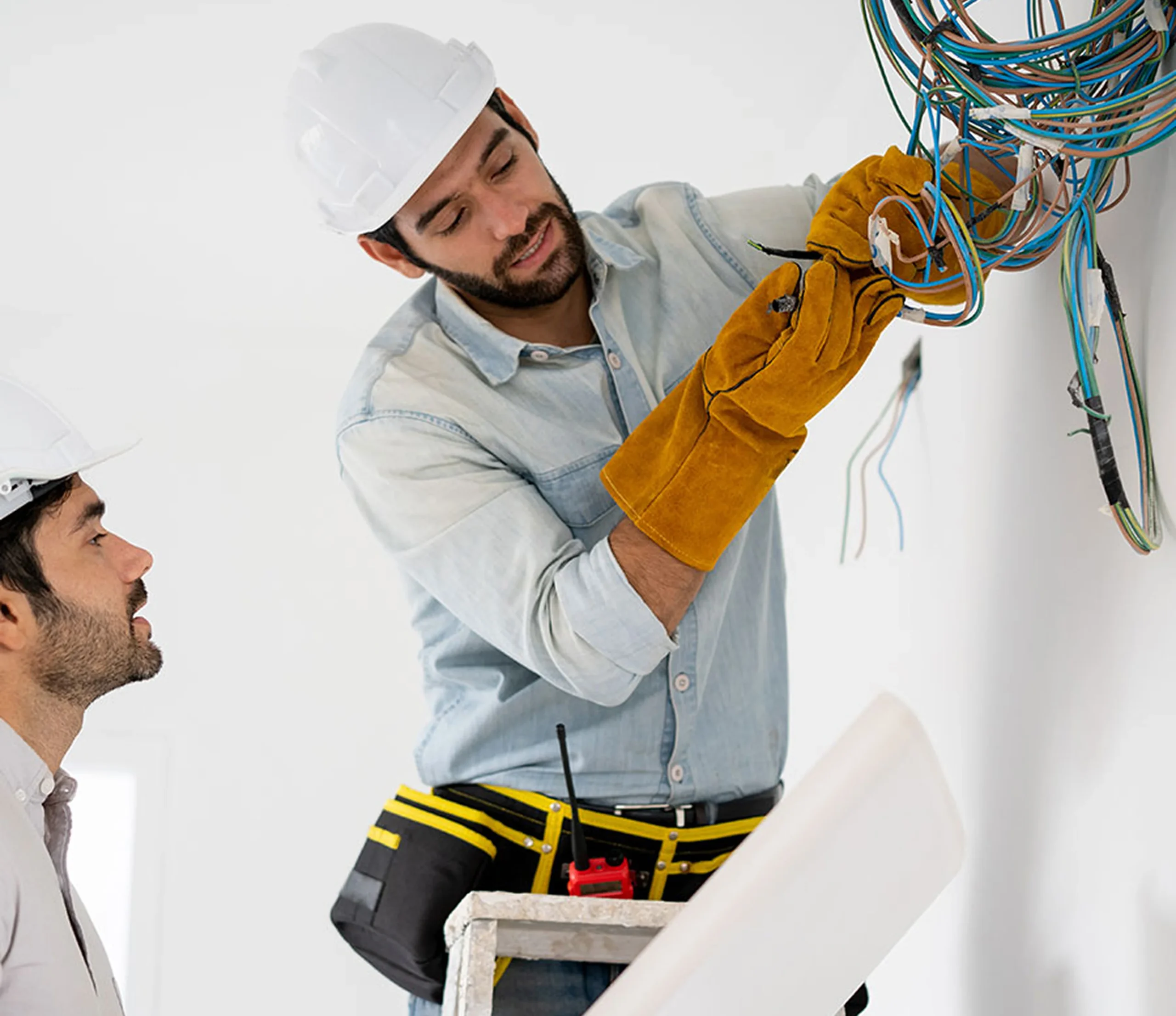 Two men wearing hard hats work on exposed electrical wiring; one is connecting wires while wearing protective gloves, the other observes. Two men wearing hard hats work on exposed electrical wiring; one is connecting wires while wearing protective gloves, the other observes.